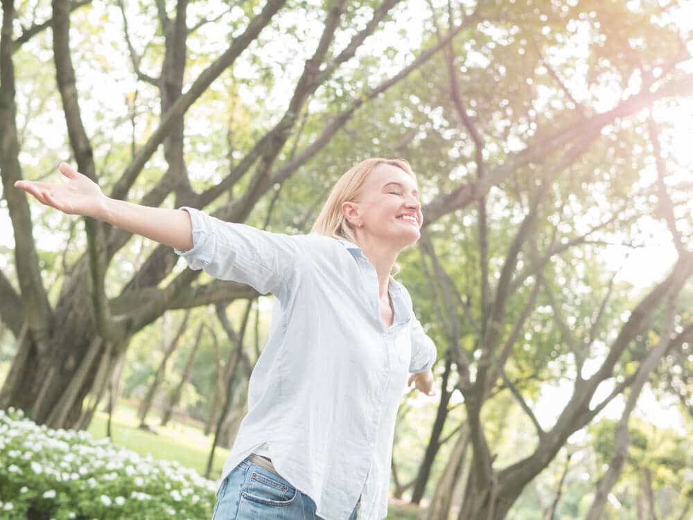 A middle-aged woman with her arms outstretched, her eyes closed, and a large smile on her face as she enjoys the nature. 