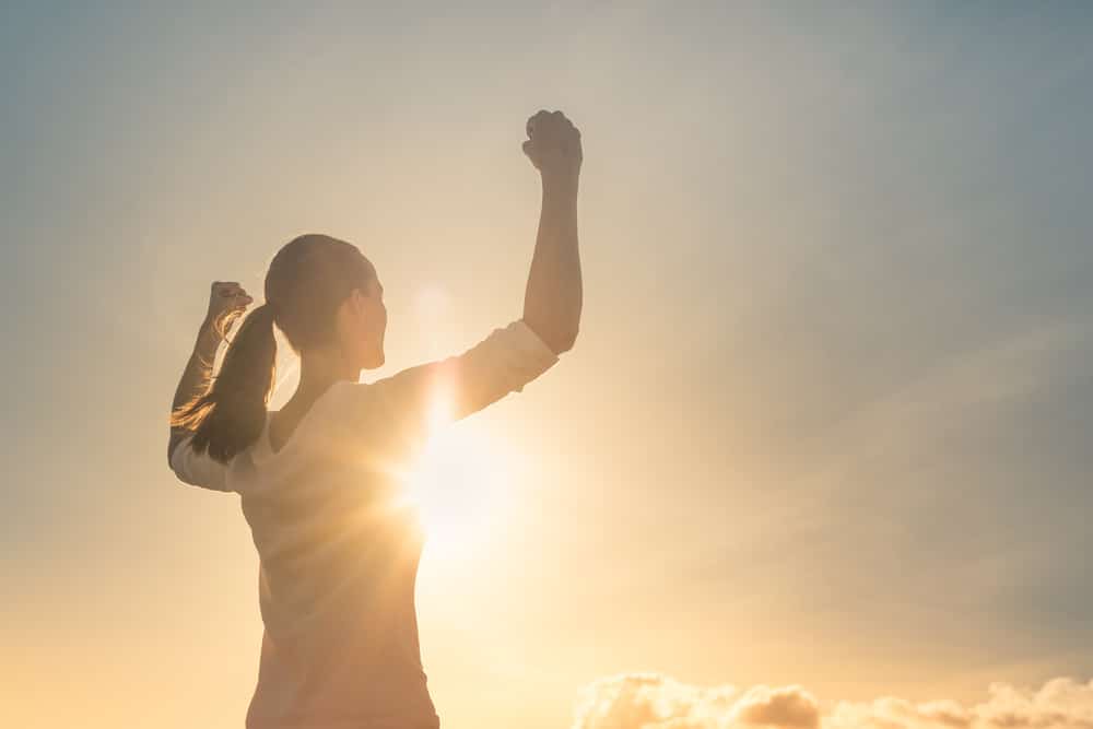 Woman facing the sun with both arms raised, posing with power, determination, and motivation. 