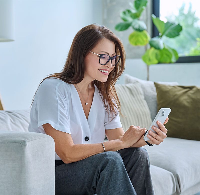 Woman with glasses smiling while using her phone on a sofa